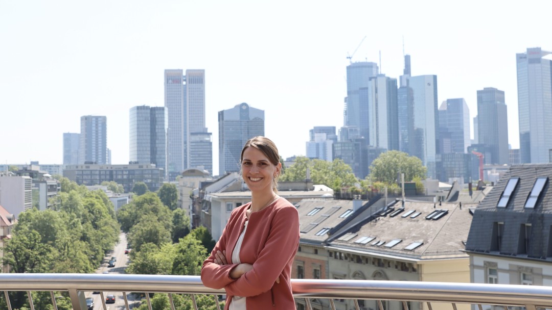 Lisa Hartnagel, Mitarbeiterin des Rechtsbereichs der KfW. Sie trägt ein weißes Oberteil und einen hellroten Blazer. Hinter ihr ist die Skyline von Frankfurt zu sehen.