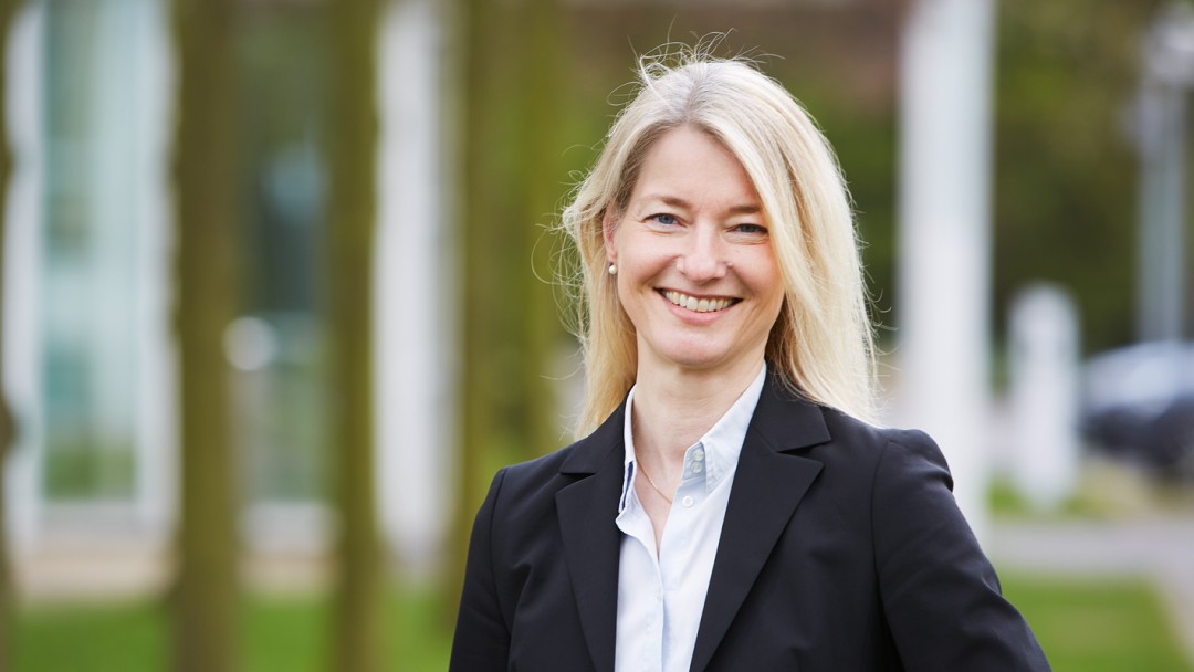Tanja Werner, Mitarbeiterin des Rechtsbereichs der KfW, steht vor einem KfW Gebäude mit Grünfläche. Sie trägt eine weiße Bluse und schwarzen Blazer.