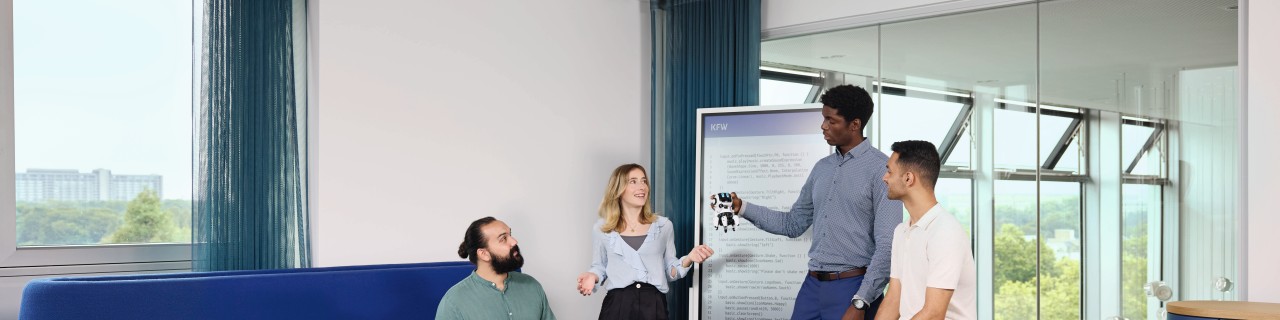 Eine diverse Gruppe (Personen verschiedenen Geschlechts, Alters und Herkunft) befindet sich in einem Besprechungsraum und arbeitet zusammen an der Programmierung von kleinen Robotern. Im Hintergrund ist ein Smartboard mit den Codes zu sehen.