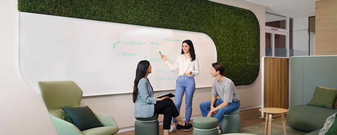 Drei junge Nachwuchskräfte befinden sich an einem Whiteboard im Digi-Café der KfW und brainstormen engagiert zusammen zum Thema hashtag Team KfW. Eine junge Frau mit dunklen mittellangen Haaren steht dabei mit einem Stift an dem Board, welches mit einer grünen dicht bewachsenen Mooswand umrandet ist. Eine weitere junge Frau mit dunklen langen Haaren sowie ein junger Mann mit braunen Haaren sitzen auf Hockern daneben und besprechen die Ergebnisse. Sie hält ein Tablet in der Hand und gleicht die Punkte mit ihren Notizen ab.