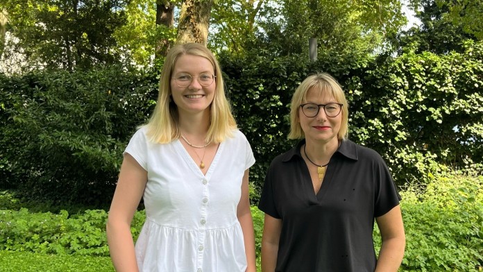 Gruppenbild von Luisa Struck, KfW Mitarbeiterin mit dem Zuständigkeitsbereich Inklusion und Erika Holz, Inklusionsbeauftragte der KfW. Luisa Struck hat mittellange blonde Haare, trägt eine Brille mit hellem Rahmen und eine weiße T-Shirtbluse. Erika Holz hat kinnlange blonde Haare, trägt eine Brille mit dunklem Rahmen und eine schwarze T-Shirtbluse. Beide stehen im begrünten Innenhof der KfW, im Hintergrund sind Büsche und Bäume zu sehen.