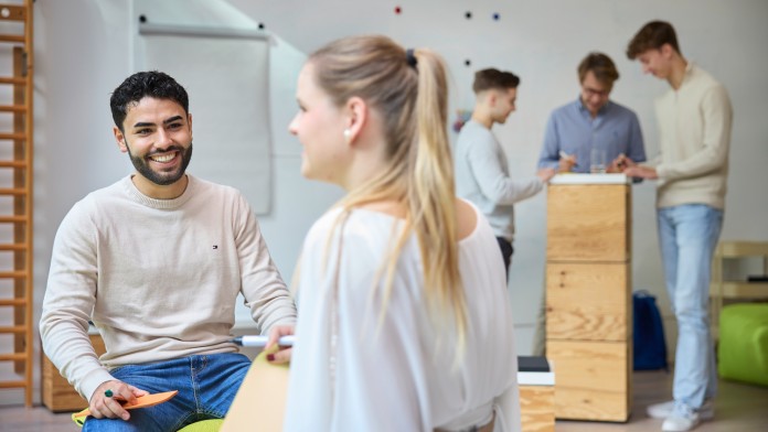 Es befinden sich fünf Nachwuchskräfte in einem kreativen Arbeitsraum der KfW. Zwei sitzen auf einer Bank und diskutieren angeregt, dabei halten Sie Karteikarten in den Händen. Im Hintergrund stehen drei weitere Nachwuchskräfte, die ebenfalls etwas ausarbeiten.