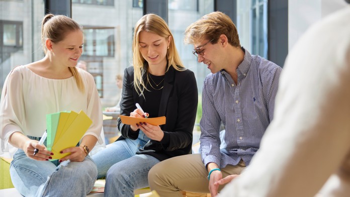 Drei Nachwuchskräfte sitzen auf einer Bank und brainstormen zum Thema Zusammenarbeit. Die mittlere Nachwuchskraft leitet das Gespräch und notiert das ausgearbeitete Ergebnis auf einer orangen Karteikarte.