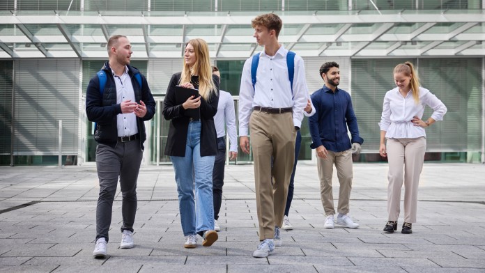 Eine diverse Gruppe von sieben Nachwuchskräften bewegt sich von einem modernen KfW Gebäude weg. Die Personen sind Business Casual angezogen, einige tragen Rucksäcke und sie unterhalten sich.