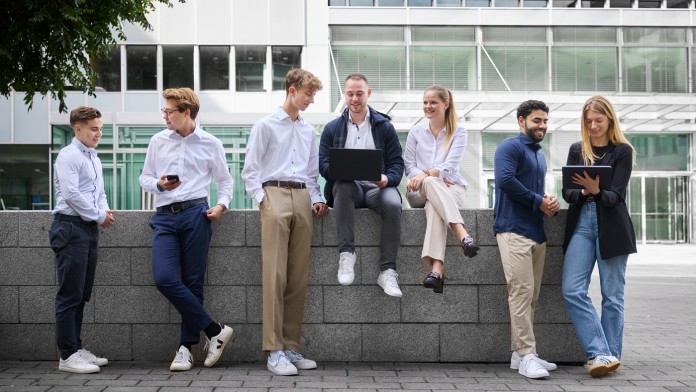 Eine diverse Gruppe von sieben Nachwuchskräften befindet sich vor einem modernen KfW Gebäude. Die Personen sind Business Casual angezogen. Manche stehen, andere sitzen auf einer niedrigen Mauer. Sie tauschen sich aus und schauen dabei auf Tablets oder Laptops.