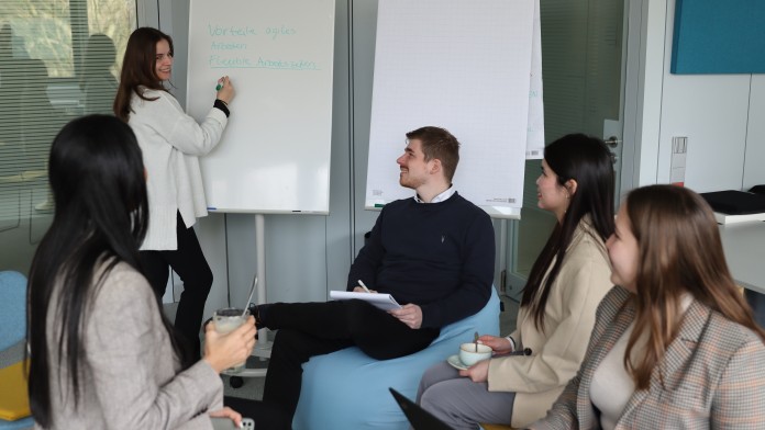 Eine Gruppe von diversen Nachwuchskräften befindet sich in einem modernen Arbeitsraum der KfW. Sie brainstormen zum Thema Vorteile von agilem Arbeiten.