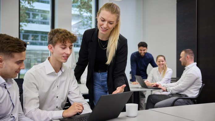 Drei Nachwuchskräfte, die sich im vorderen Bereich des Bildes befindet, sind in einem modernen Seminarraum und betrachten eine gemeinsame Präsentation auf einem Laptop. Im Hintergrund besprechen drei weitere Personen angeregt ihre Arbeitsergebnisse.