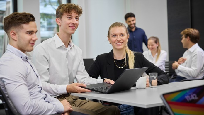 Eine diverse Gruppe von Nachwuchskräften, die an zwei Tischen verteilt ist, bespricht in kleinen Gruppen die nächsten Projekte. Die drei Personen im vorderen Teil des Bildes hören gespannt ihrem Arbeitskollegen zu.