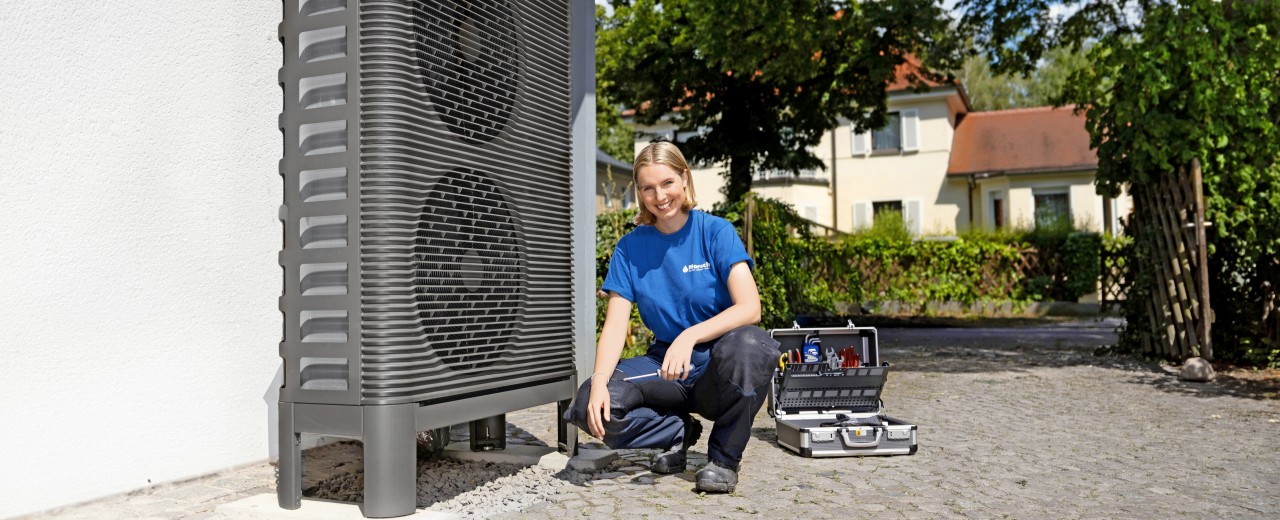 Die Anlagenmechanikerin kniet vor einer Wärmepumpe mit Werkzeug in der Hand.